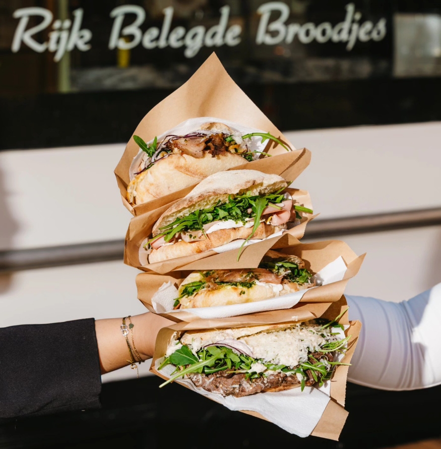 Hand houdt vier rijk belegde Italiaanse broodjes in papier vast voor de winkel